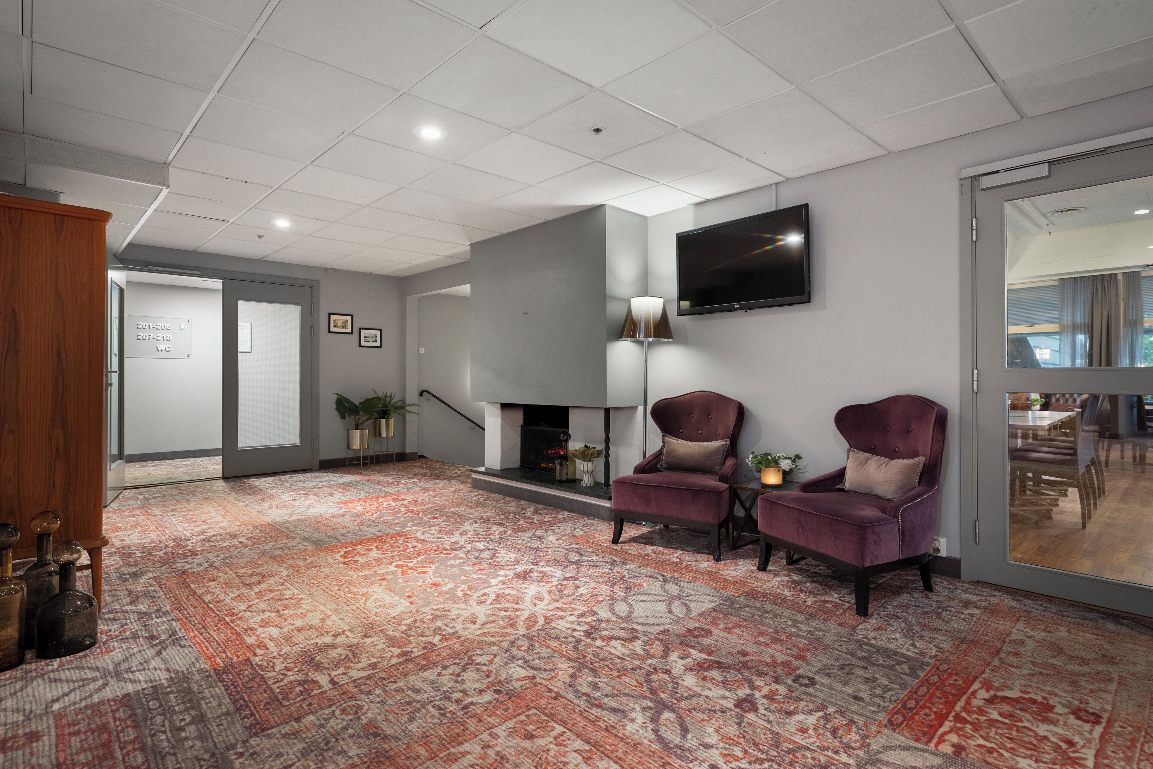 Eine moderne Lobby mit Teppichmuster, zwei Sesseln, einem Kamin und einem an der Wand befestigten Flachbildfernseher.