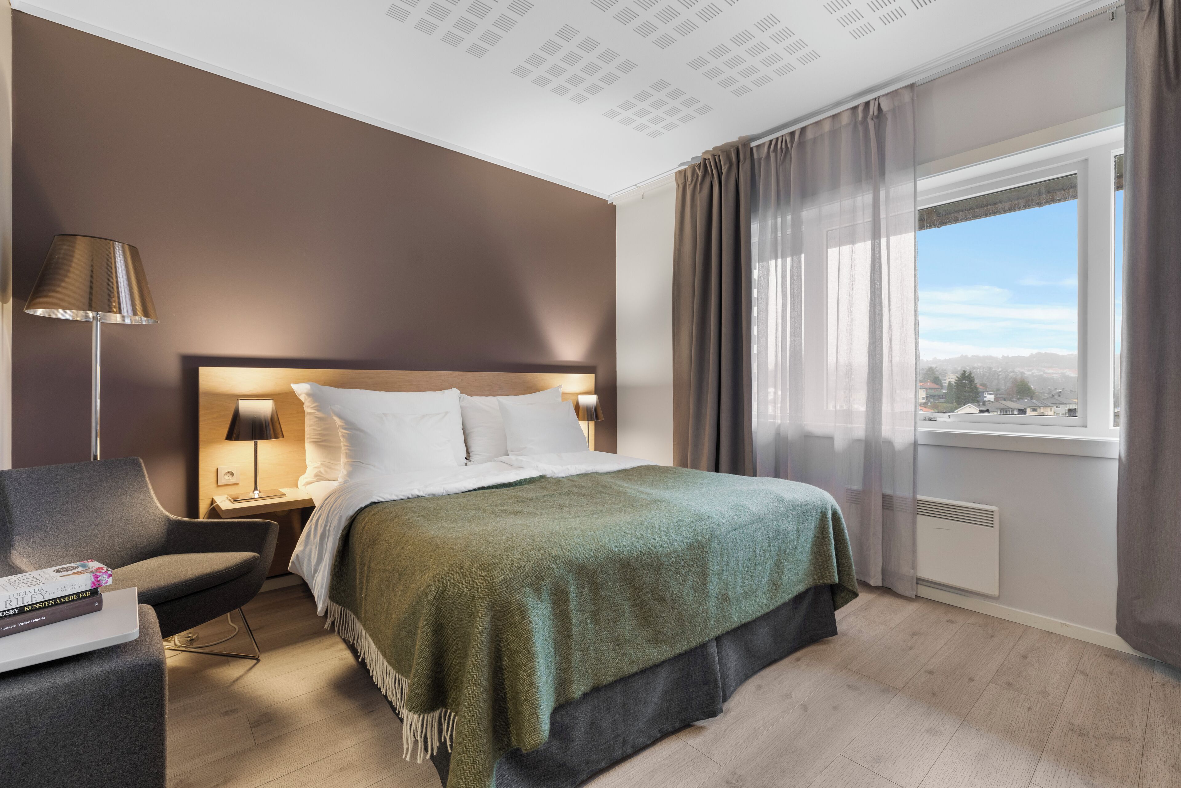 Una habitación de hotel moderna con una cama grande, un escritorio con lámpara, una silla y vistas de la ciudad a través de un ventanal amplio.