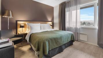 Una habitación de hotel moderna con una cama grande, un escritorio con lámpara, una silla y vistas de la ciudad a través de un ventanal amplio.