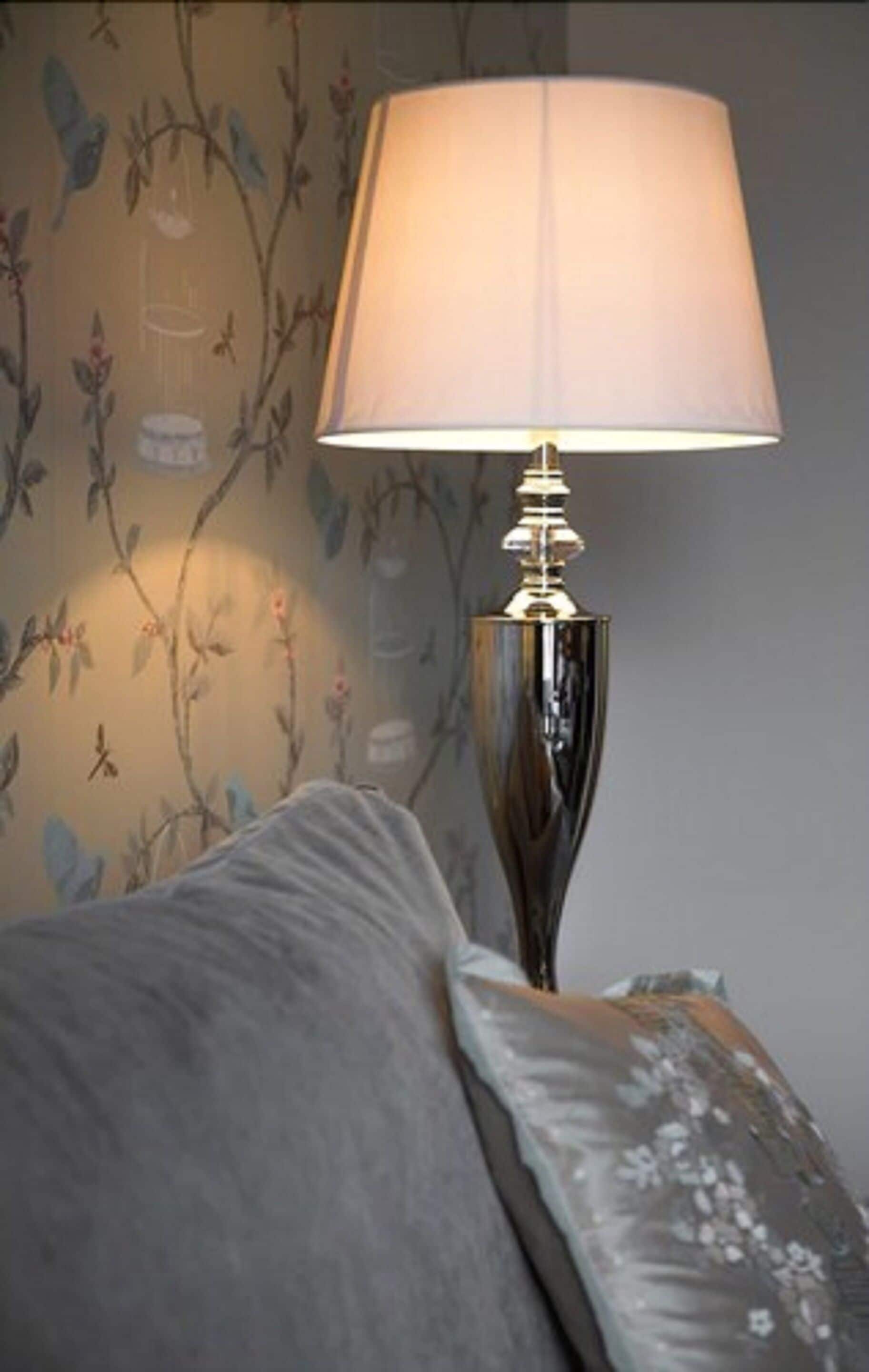 Una lámpara con pantalla color beige sobre una mesita de noche junto a una cama con ropa de cama estampada.