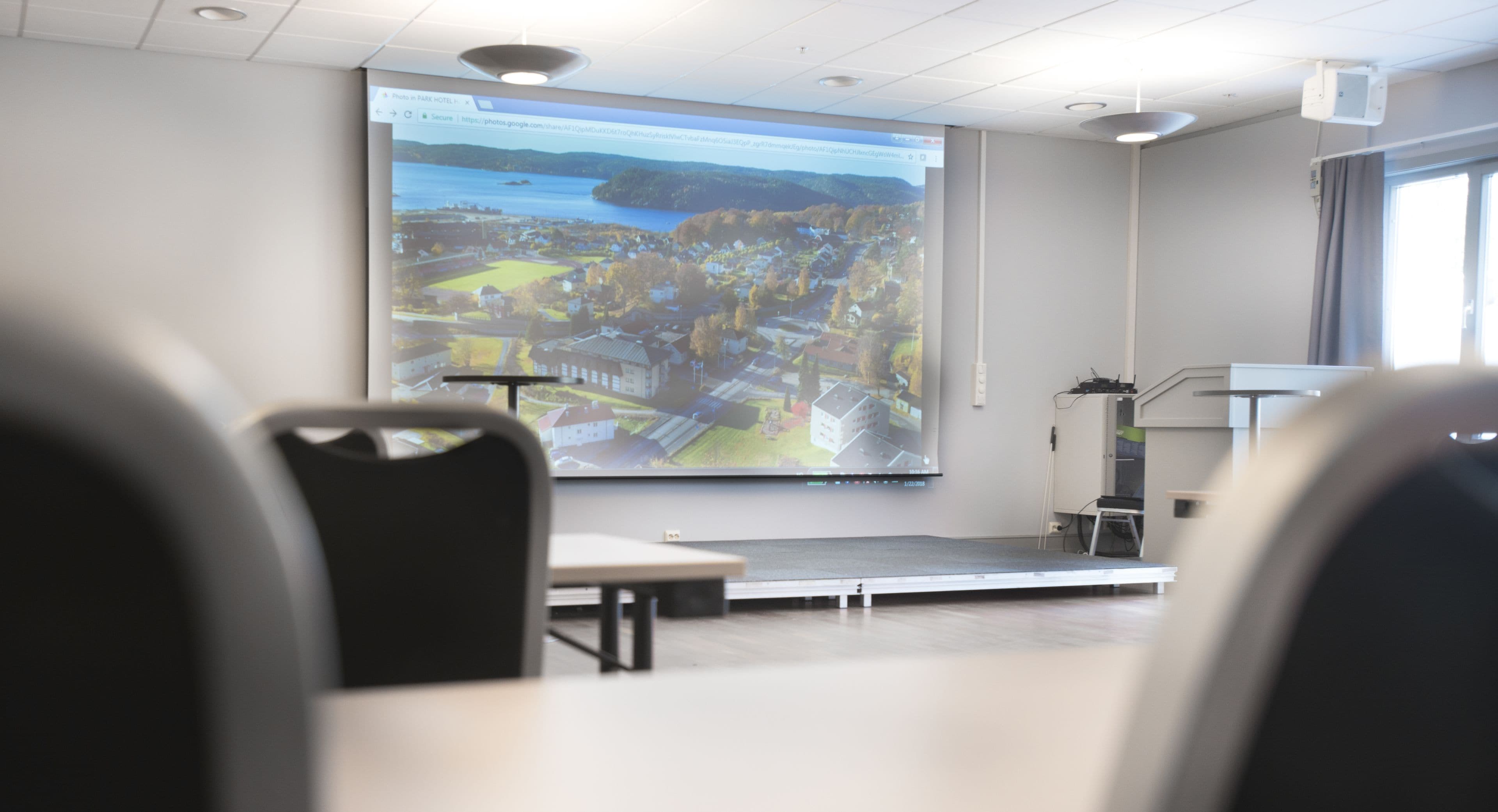 A classroom with a large projection screen displaying a scenic landscape, a podium, and a few chairs.