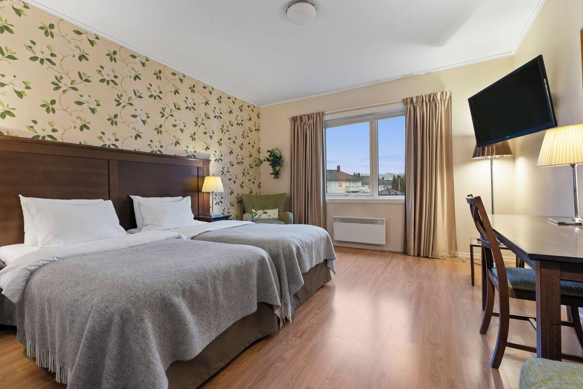 A hotel room with two beds, a TV, a desk with a chair, a window with curtains, and a view of buildings outside.