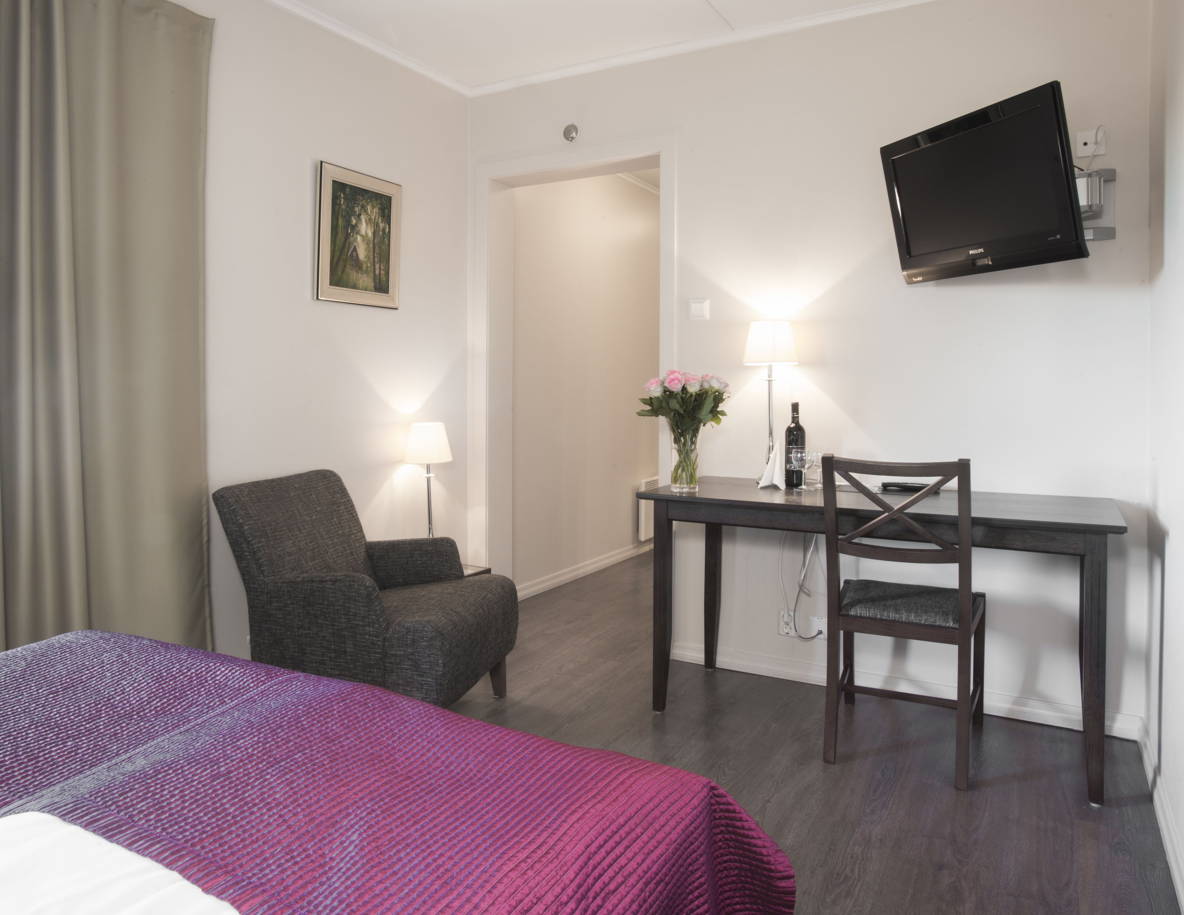 A hotel room with a bed, a desk with a chair, a TV mounted on the wall, a lamp, and a vase with flowers.
