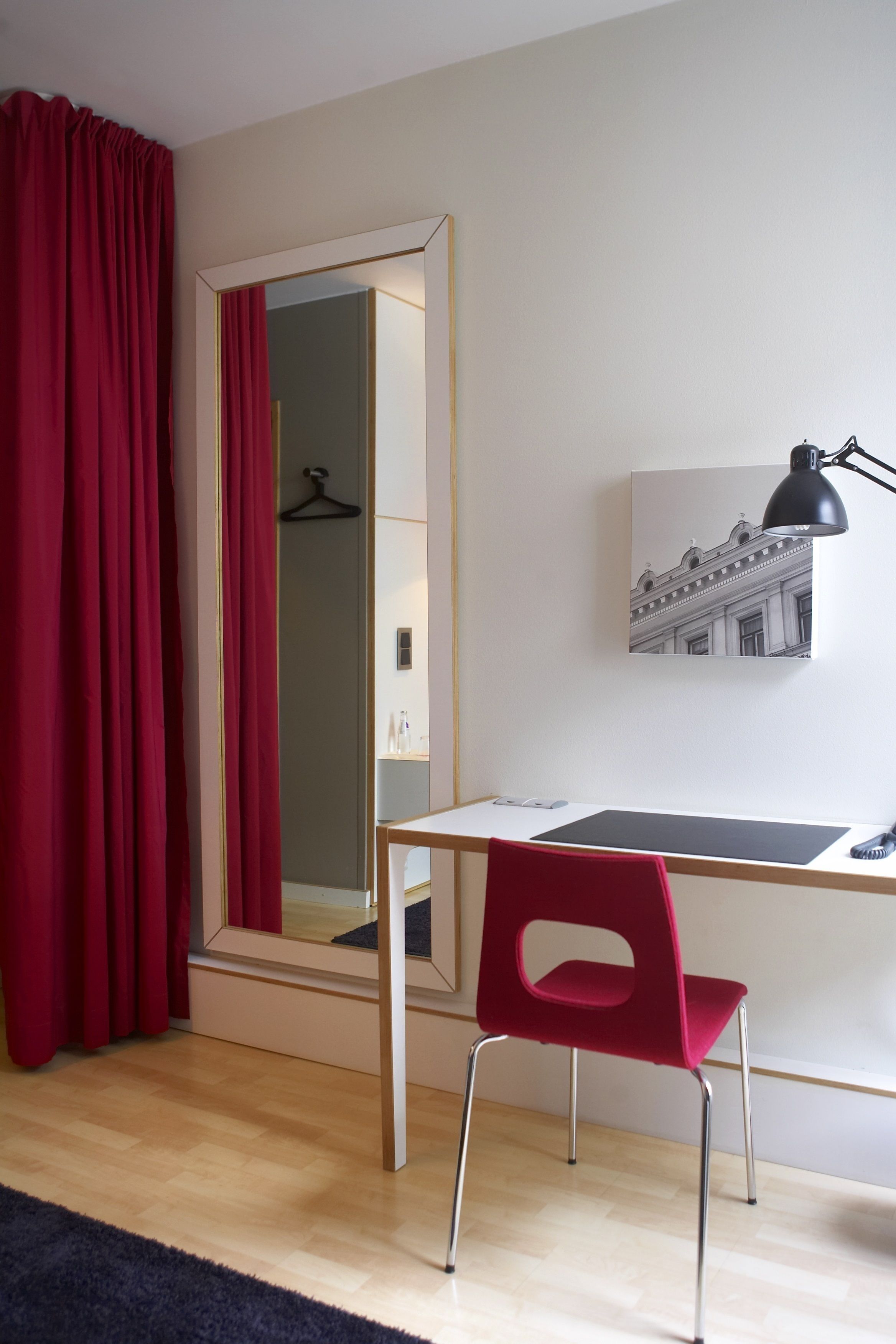 A room with a wooden desk, a red chair, a black desk lamp, a framed picture on the wall, and a mirror reflecting a red curtain and a hanger.