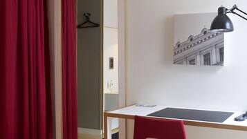 A room with a wooden desk, a red chair, a black desk lamp, a framed picture on the wall, and a mirror reflecting a red curtain and a hanger.