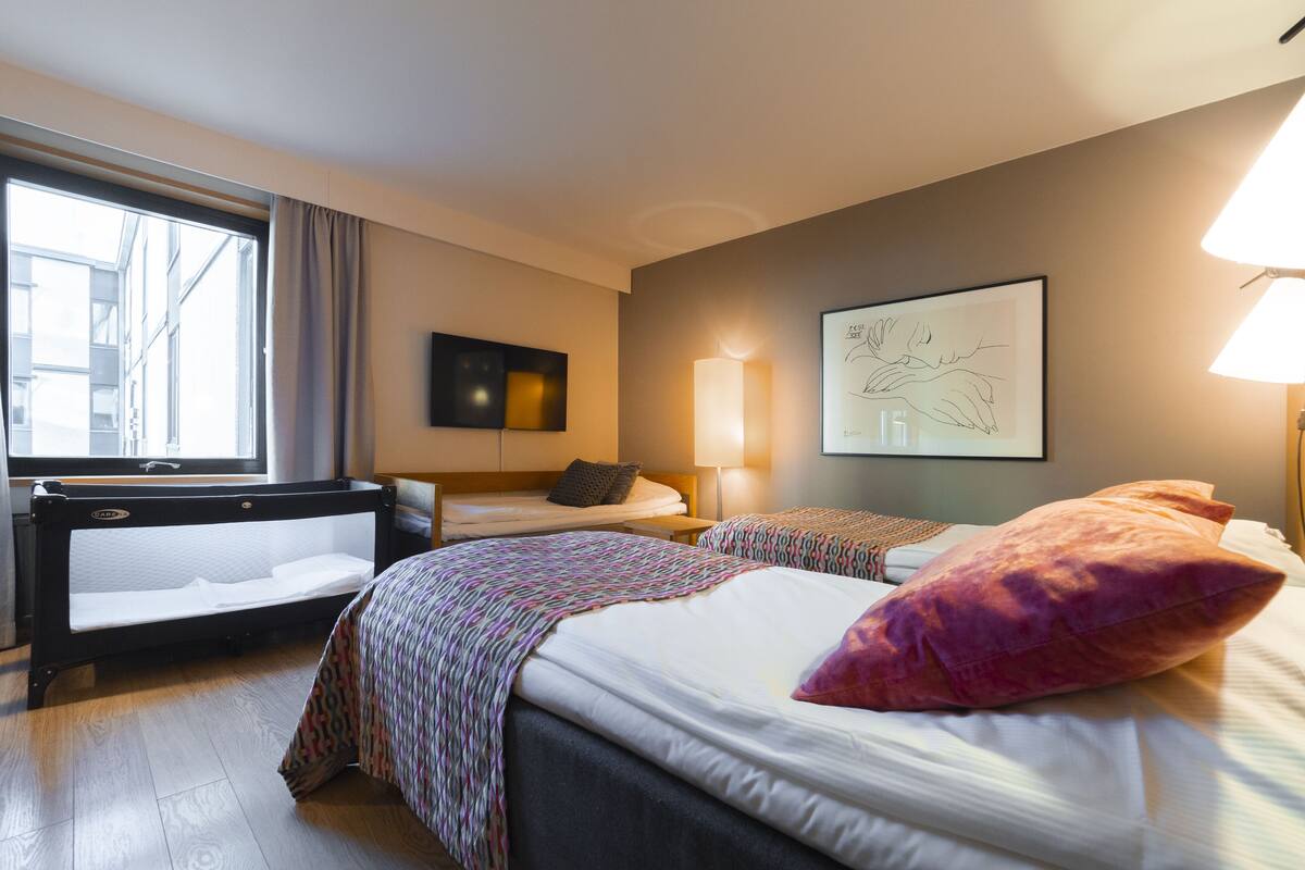 A hotel room with two beds, a desk, a chair, a window, a lamp, and a wall art piece.