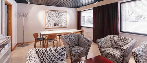 A modern room with a wooden ceiling, a dining area with patterned chairs, a large window with curtains, and a framed artwork on the wall.