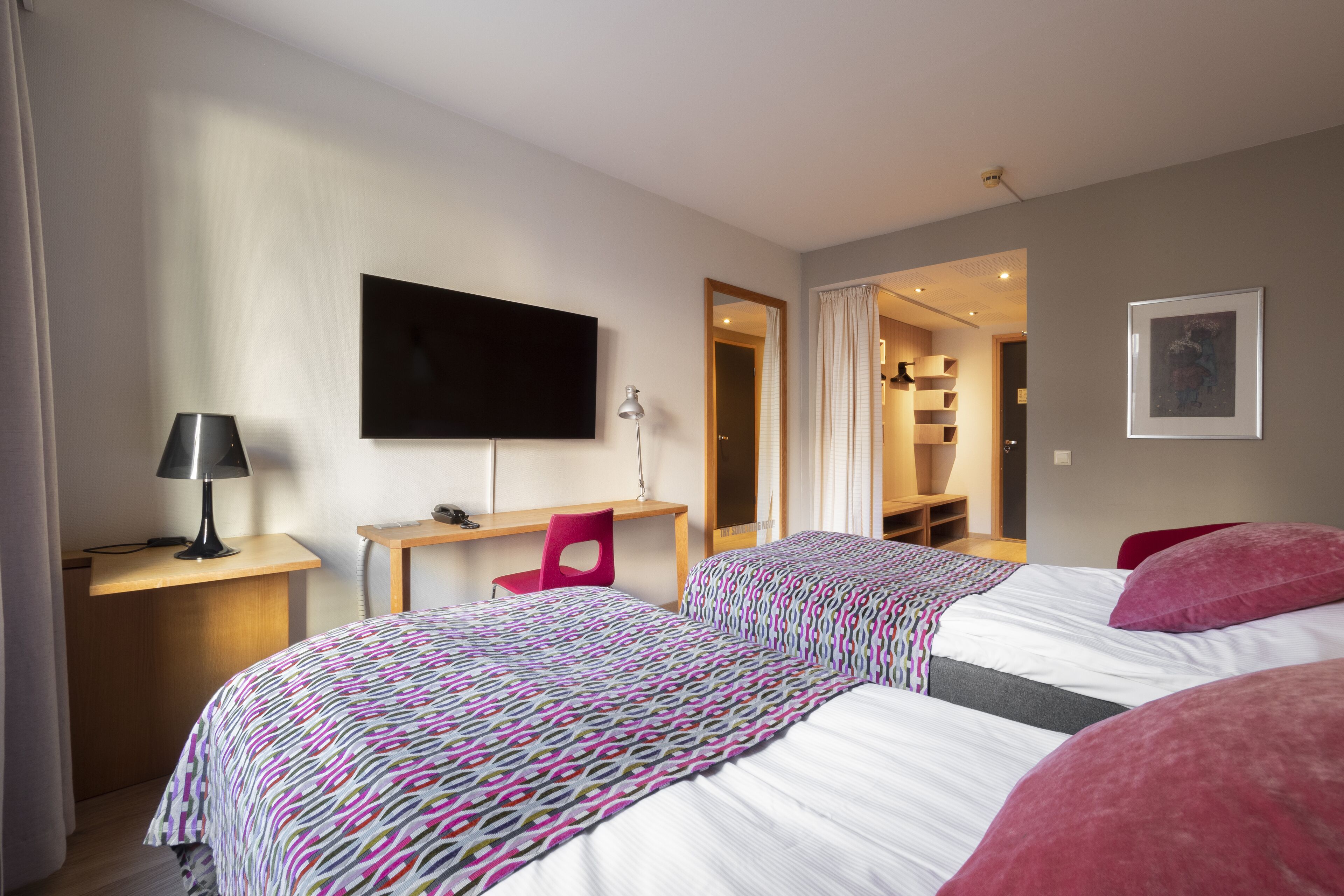 Hotellihuone, jossa on kaksi sänkyä, työpöytä, tuoli, televisio ja seinällä oleva kehystetty kuva.