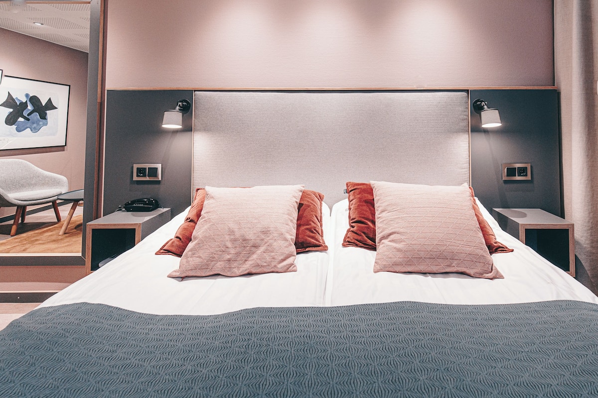 A neatly made bed with two pillows, a bedside table with a lamp, and a chair in a modern hotel room.