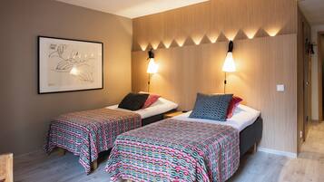 A hotel room with two beds, each with patterned bedding, a framed artwork on the wall, and wall-mounted lighting.
