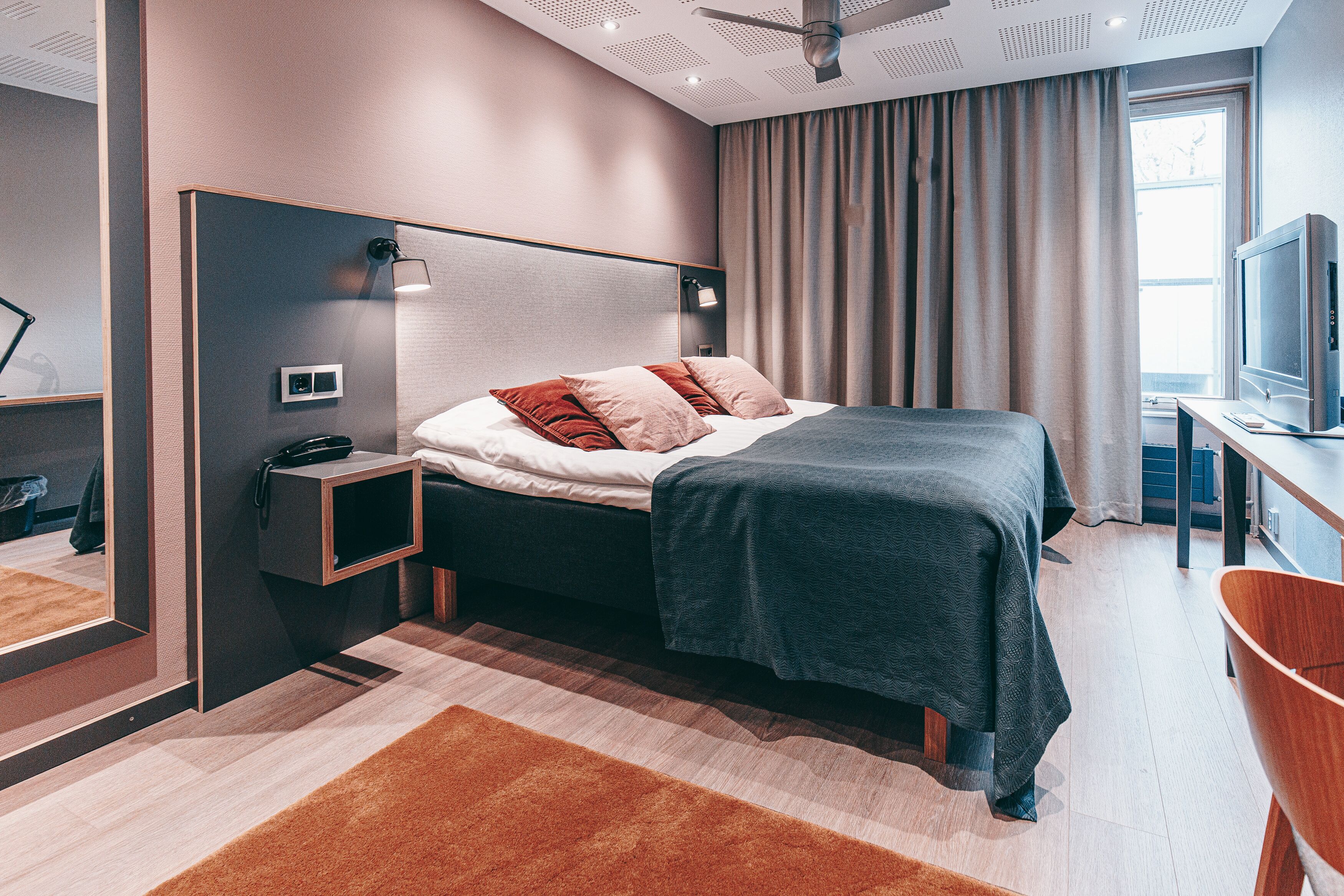 A modern hotel room with a bed, a desk with a television, a mirror, and a window with curtains.