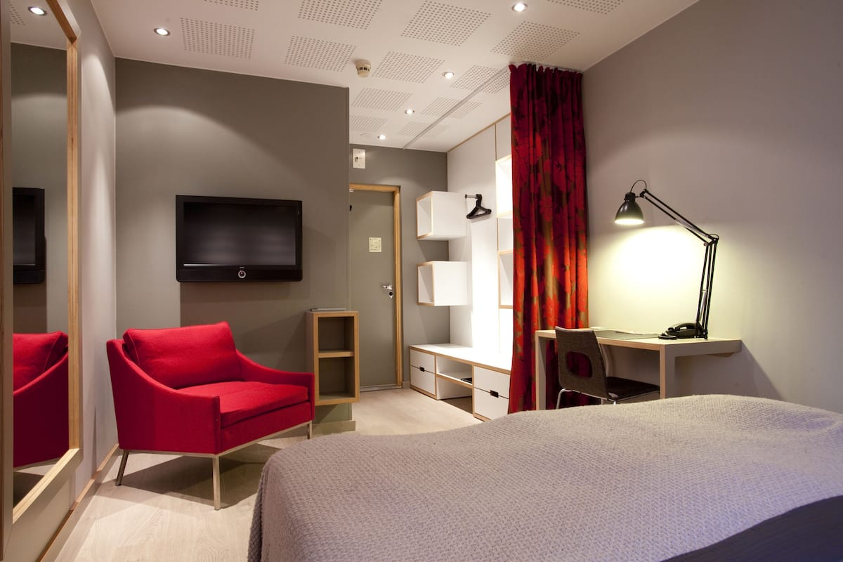 A hotel room with a bed, a red chair, a desk with a lamp, a TV, and a wardrobe.