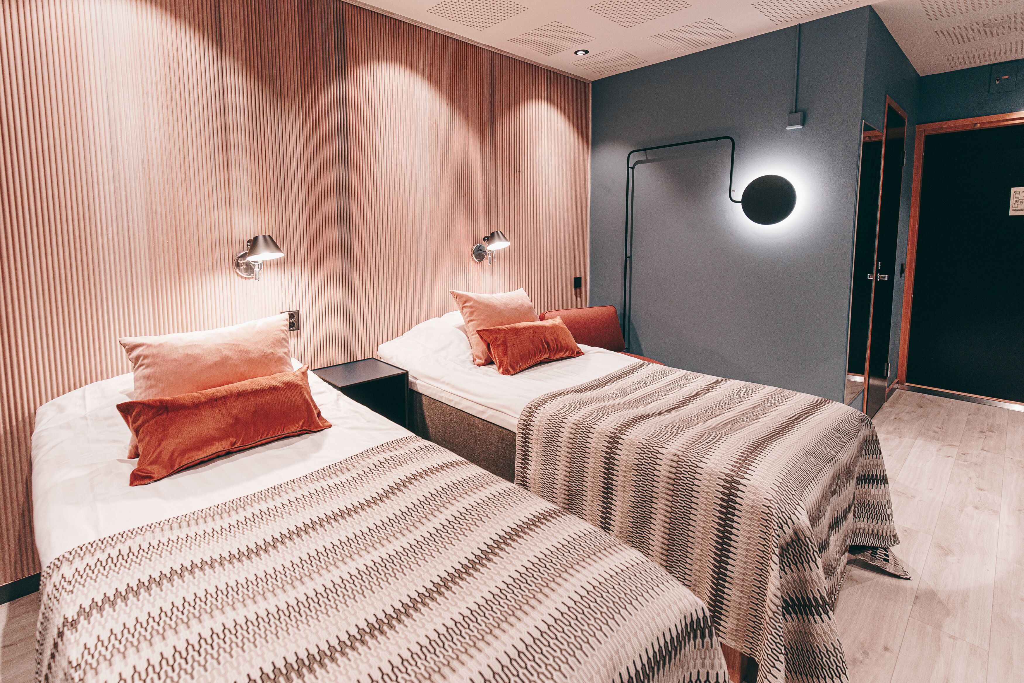 Une chambre d’hôtel avec deux lits, chacun agrémenté d’une couvre-lit à motifs et de coussins, d’une table de chevet et d’une lampe fixée au mur.