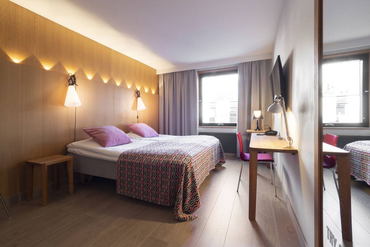 A modern hotel room with a wooden headboard, a bed with patterned bedding, a desk with a chair, and a large window.