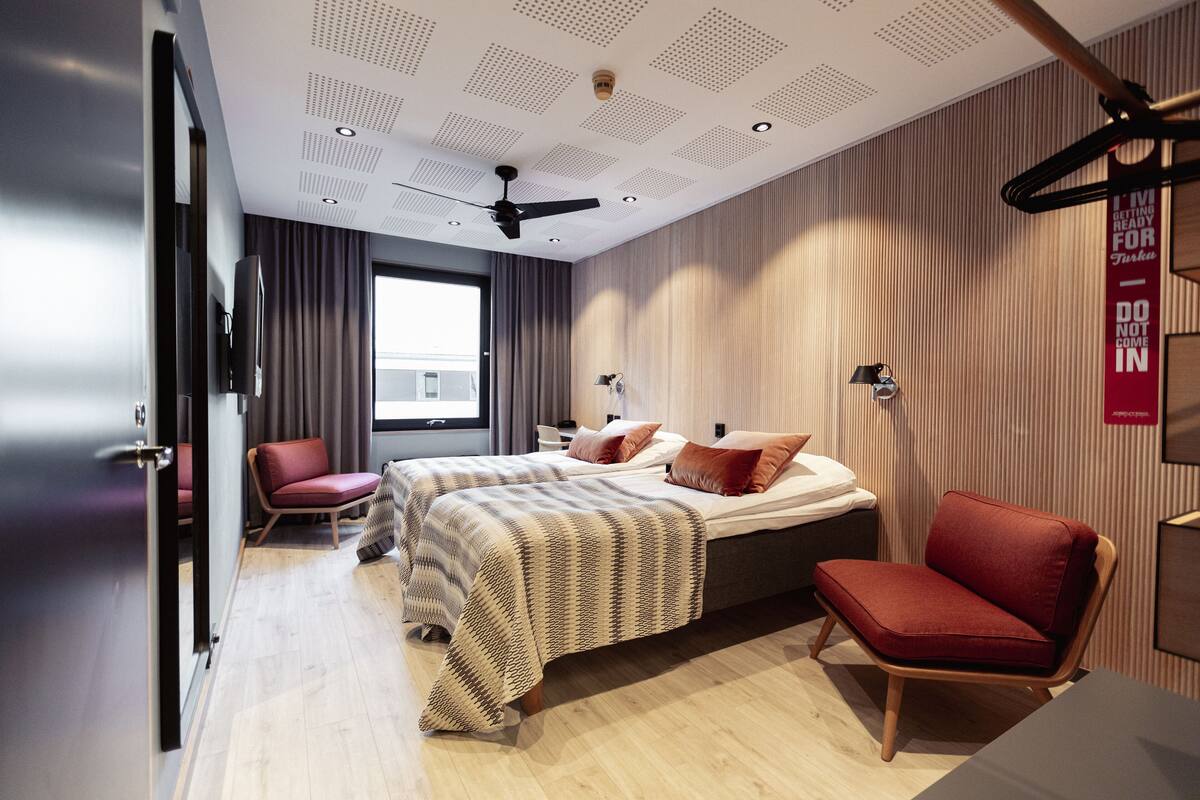 A modern hotel room with a bed, a red chair, a desk, and a large window.