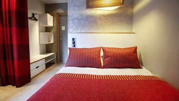 A hotel room with a bed, red curtains, a bedside table, a lamp, and a wall-mounted light fixture.