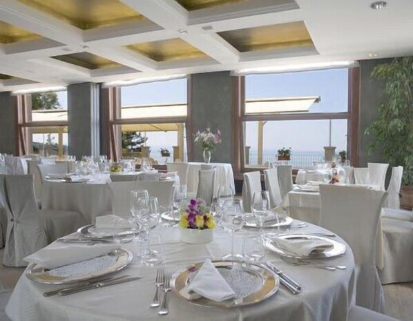 Un espace repas avec des tables rondes dressées pour un repas formel, de grandes fenêtres offrant une vue sur la mer et un design moderne au niveau du plafond.
