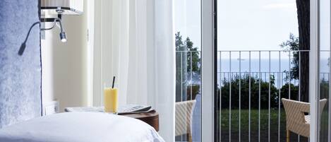 Habitación de hotel con cama, mesita de noche con lámpara, una silla y vistas al mar desde un balcón.