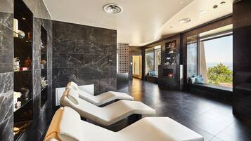 A modern spa area with dark marble walls, a seating area with white cushioned chairs, and a large window offering a view of the outdoors.