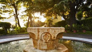 Una fuente de piedra con intrincados grabados en un estanque circular, rodeada de abundante vegetación y árboles.