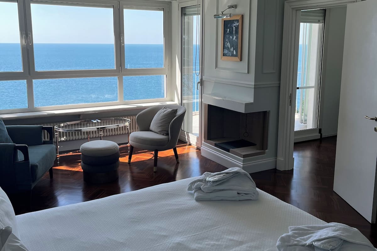 Une chambre spacieuse avec un grand lit, un coin salon comprenant un fauteuil et un pouf, une cheminée et une vue sur la mer grâce à de grandes fenêtres.
