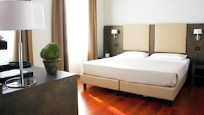 A hotel room with a bed, bedside lamps, a desk with a lamp, a potted plant, and a window with curtains.