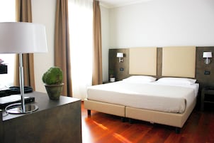 Quarto de hotel com cama, abajures, escrivaninha com luminária, planta em vaso e janela com cortinas.