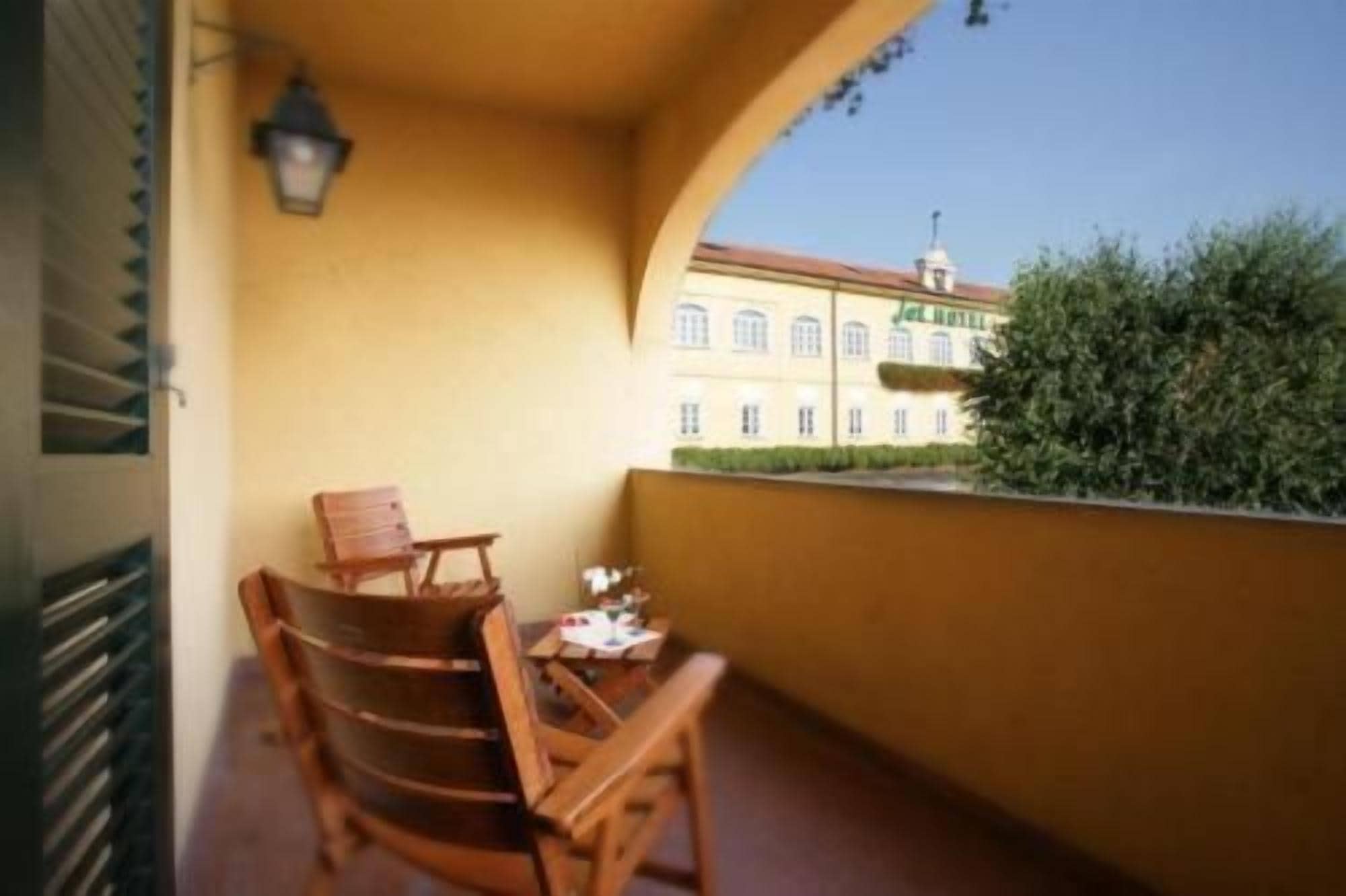 A balcony with a wooden chair and table, a lamp, and a view of a building with a red roof.