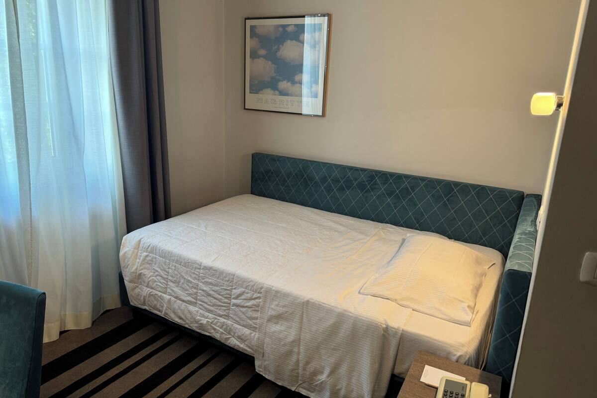 Una cama individual con una colcha blanca acolchada, cabecera con diseño de rombos, un cuadro enmarcado con nubes en la pared, y una alfombra a rayas.