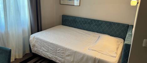 A single bed with a quilted white bedspread, a headboard with a diamond pattern, a framed picture of clouds on the wall, and a striped carpet.