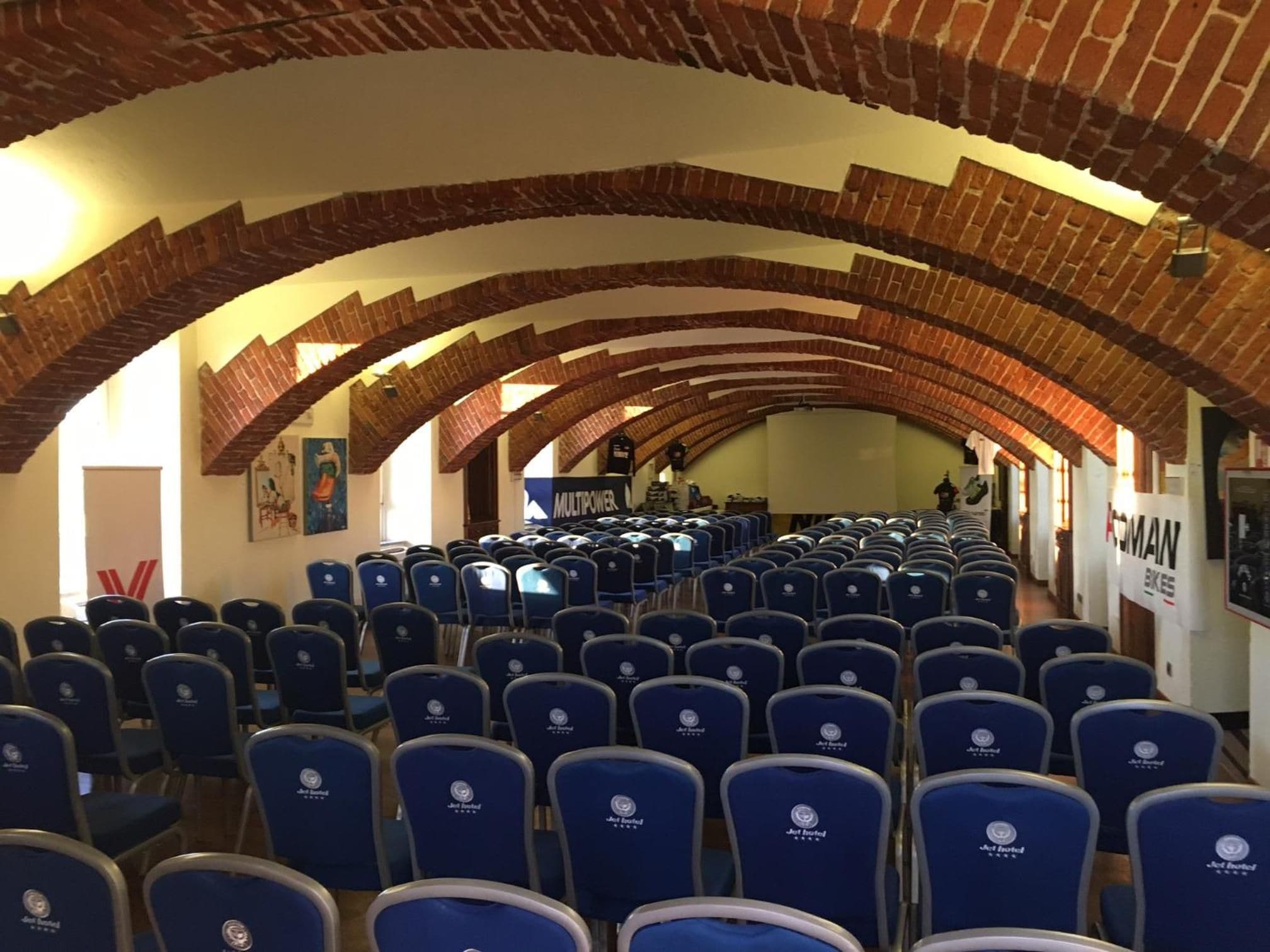 Un long plafond voûté en briques dans une salle de conférence, avec des rangées de chaises bleues.