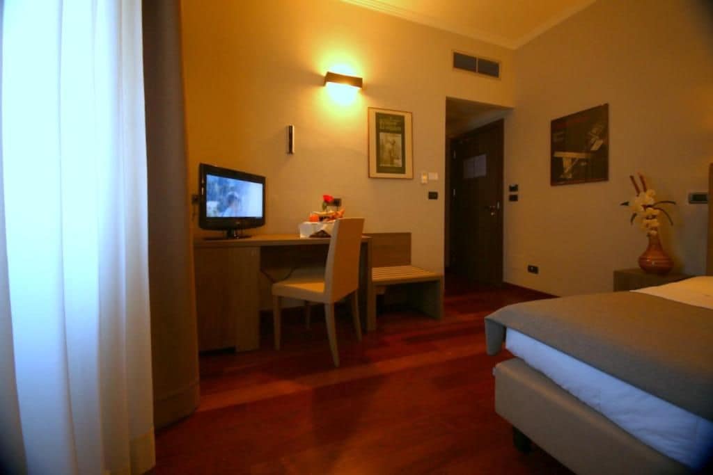 Ein Hotelzimmer mit einem Bett, einem Schreibtisch mit Stuhl, einem Fernseher und einer Blumenvase.