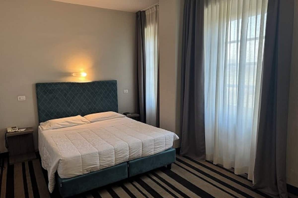 Una habitación de hotel con cama, mesita de noche, lámpara y ventana con cortinas.