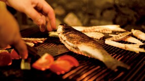 A fish grilling on a barbecue with vegetables and a hand adjusting the food.