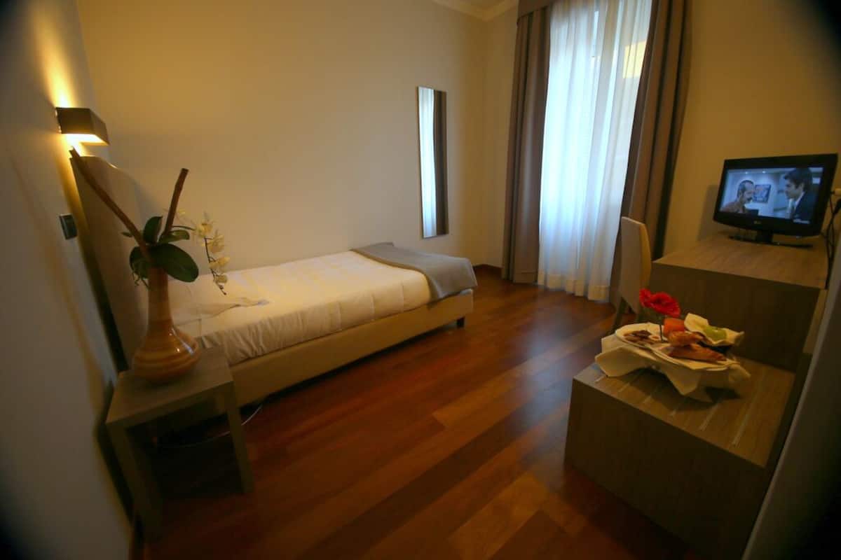 Una habitación de hotel con cama, una mesita con un florero, televisión y ventana con cortinas.