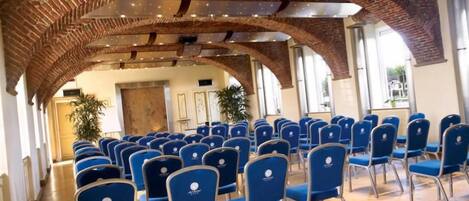 A conference room with rows of blue chairs, brick arches, and a wooden floor.