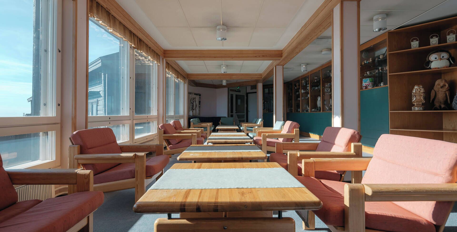 Une pièce baignée de lumière naturelle, avec des tables et des chaises en bois, de grandes fenêtres et des étagères ornées d’objets décoratifs.