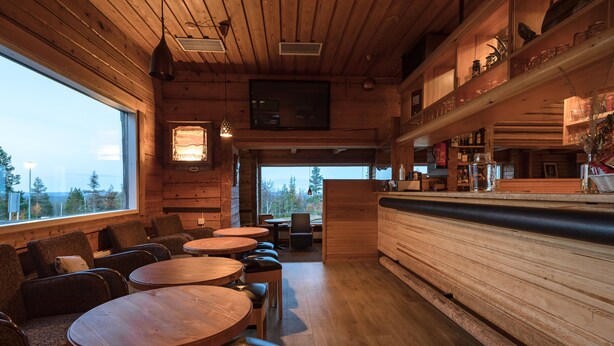 A cozy wooden interior with round tables and benches, a bar area, and a view through a large window. - Lapland Hotels Pallas (Muonio)