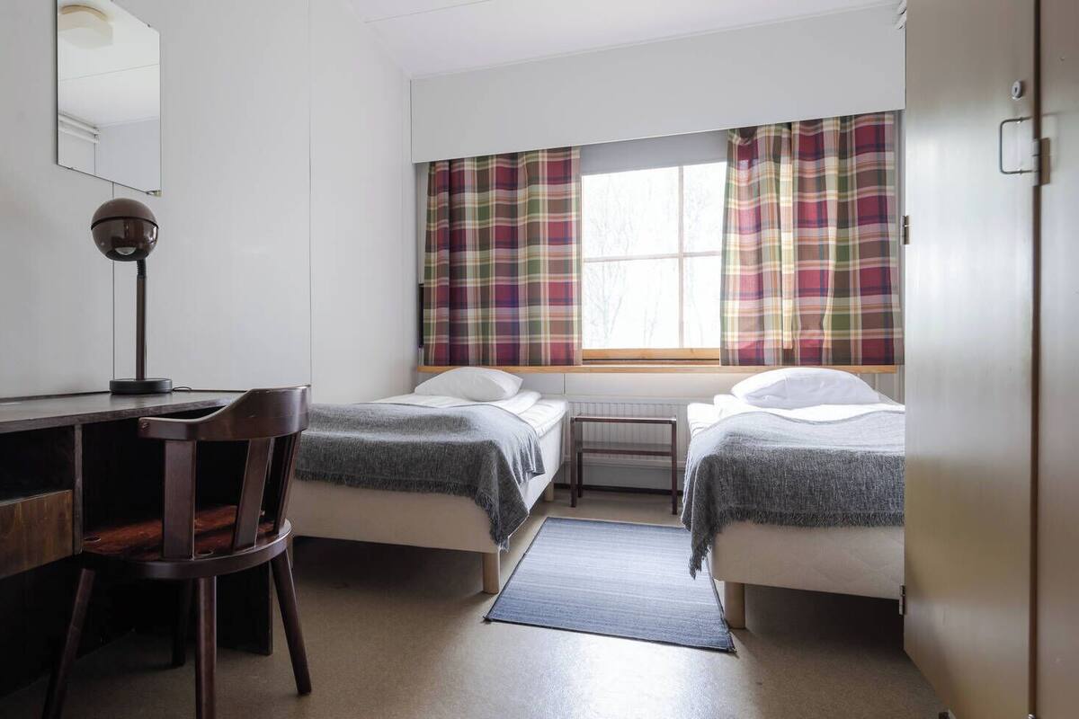 Ein Zimmer mit zwei Einzelbetten, einem Schreibtisch mit Stuhl, einem Fenster mit Karottenvorhängen und einer Heizung.