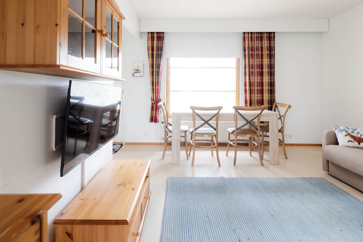Ein modernes Wohnzimmer mit Flachbildfernseher, Holzmöbeln, einem Esstisch mit Stühlen und einer Couch mit Kissen.