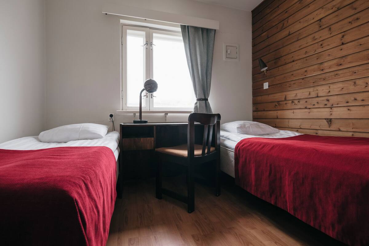 Ein Zimmer mit zwei Einzelbetten, einem Schreibtisch, einem Stuhl, einer Lampe und einem Vorhangfenster.