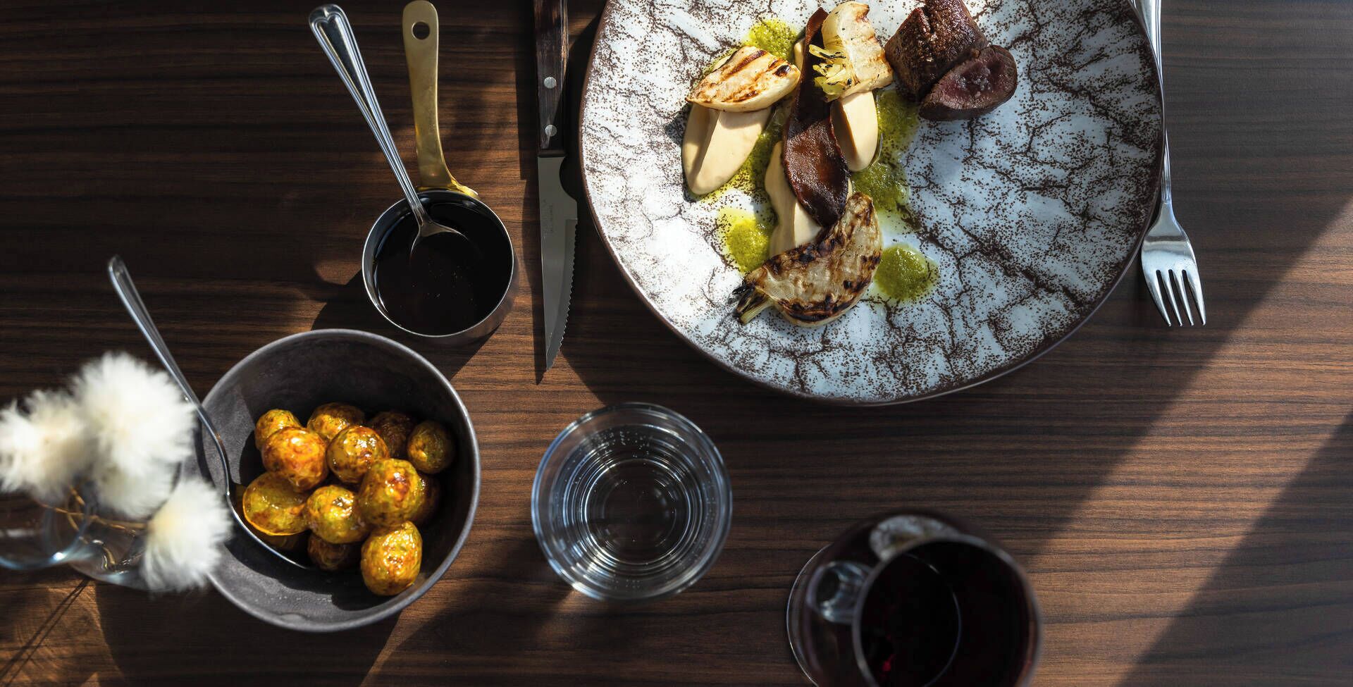 A dining table with a plate of food, a bowl of meatballs, and two glasses of red wine.