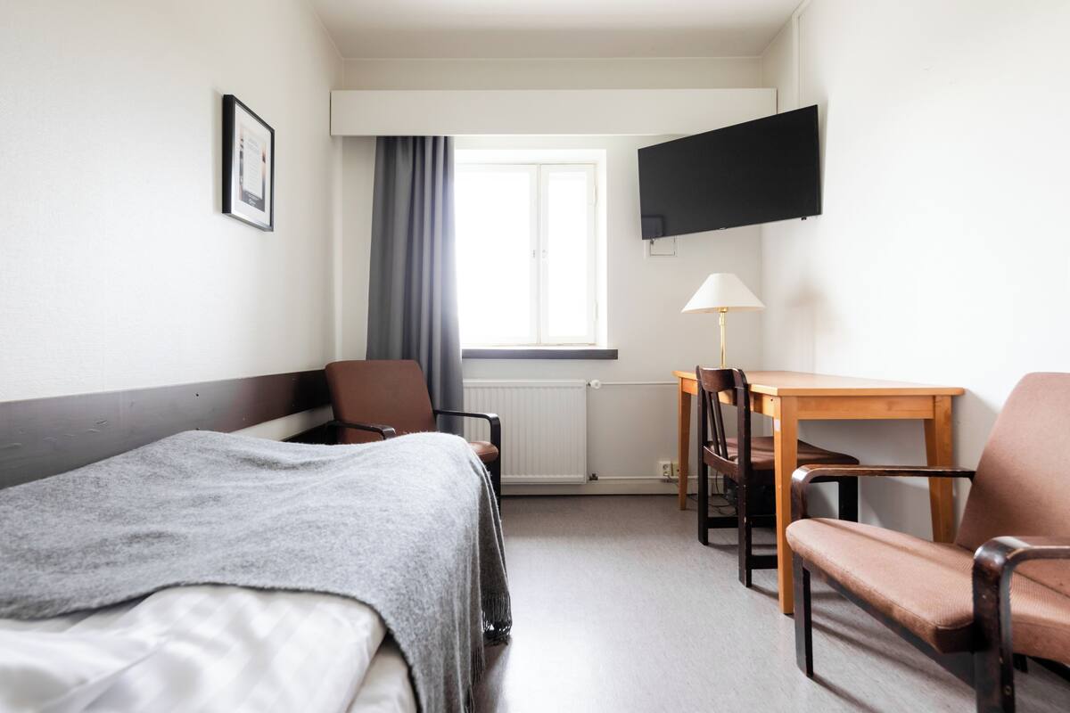 Ein Hotelzimmer mit einem Bett, zwei Sesseln, einem Tisch, einem Fernseher, einem Fenster mit Vorhängen und einer Lampe.