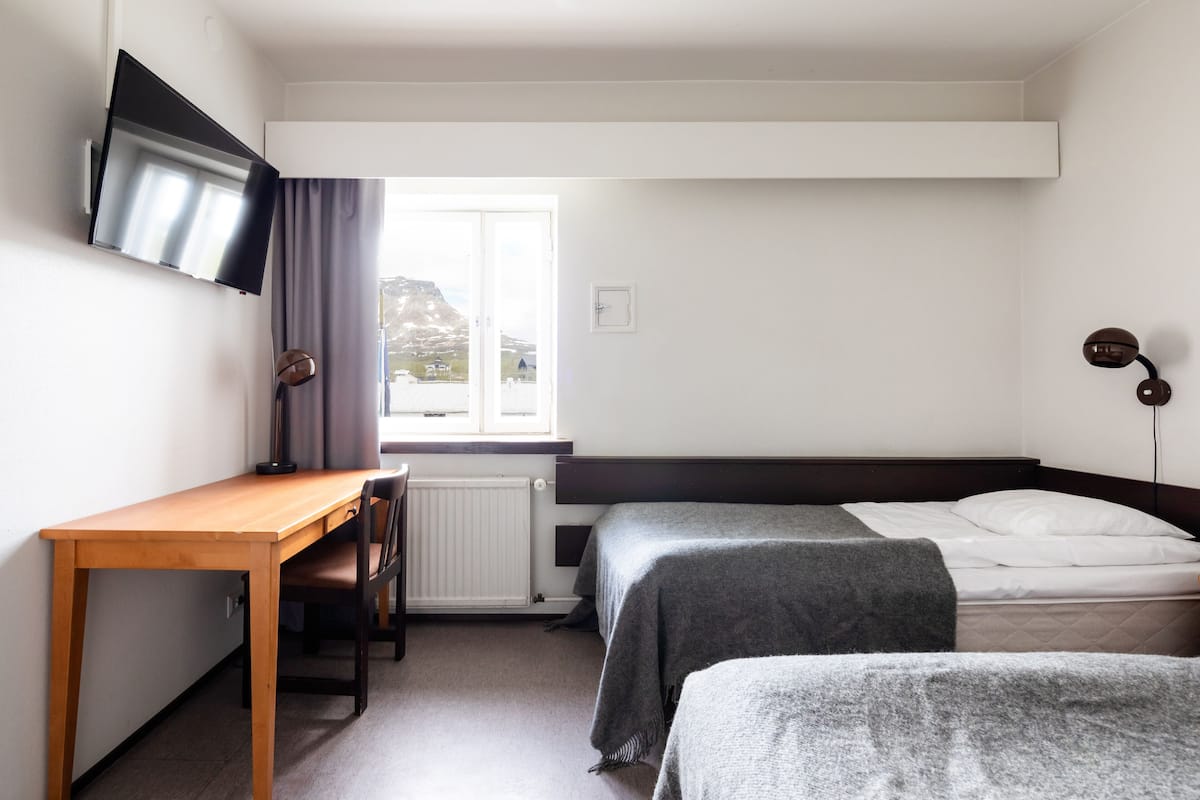 Ein Zimmer mit einem Holztisch, zwei Betten, einem an der Wand befestigten Fernseher und einem Fenster mit Vorhängen.
