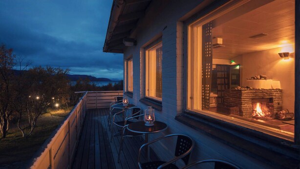 A wooden deck with outdoor seating and a view of a lit fireplace inside a building. - Lapland Hotels Kilpis (Enontekio)