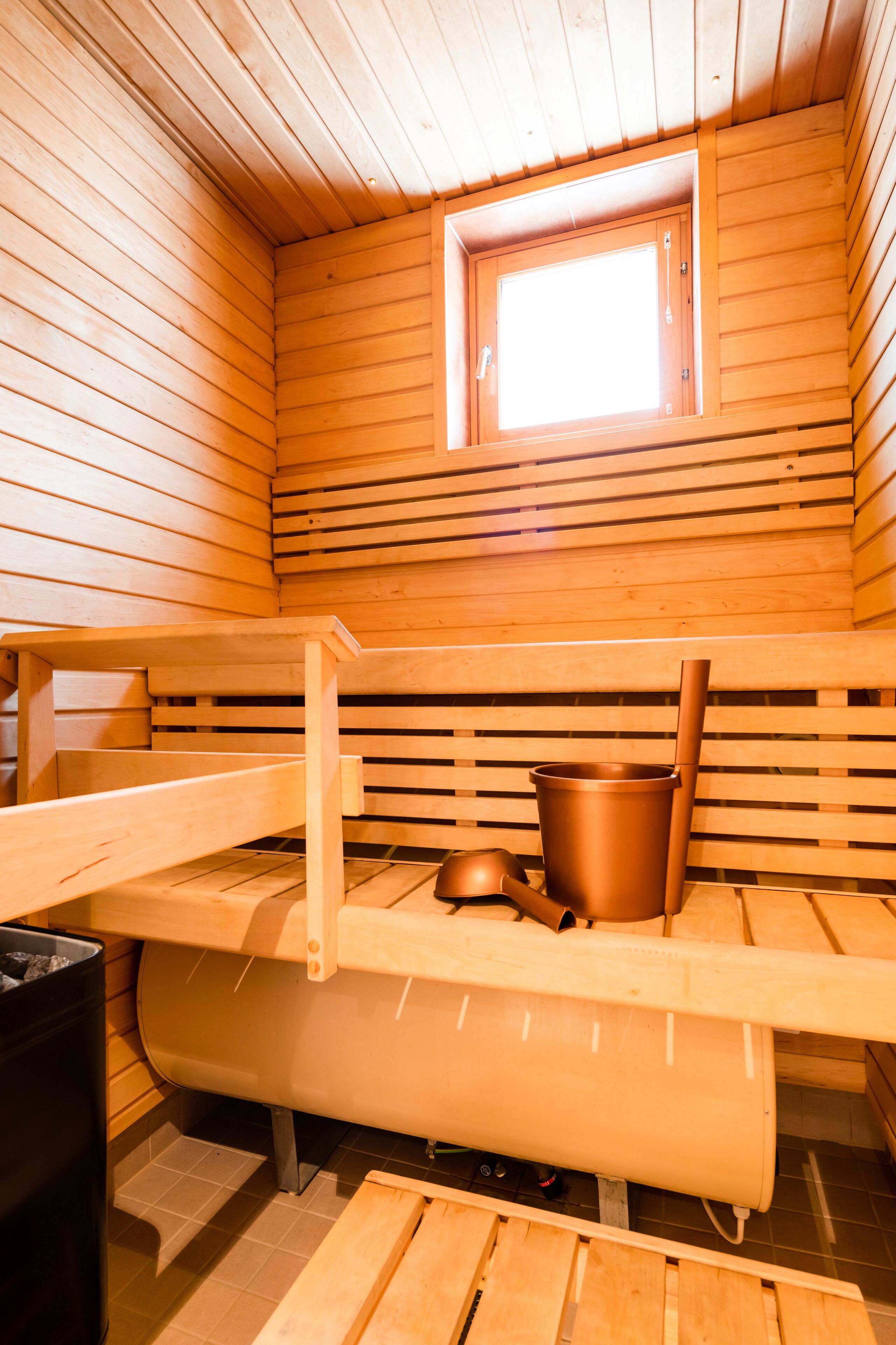 A sauna with wooden benches, a window, and a large metal container.
