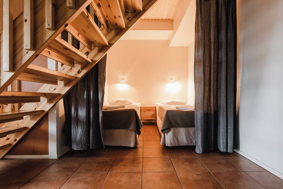 Una habitación con una escalera de madera, dos camas con ropa de cama a cuadros, una mesita de noche y lámparas fijadas a la pared.