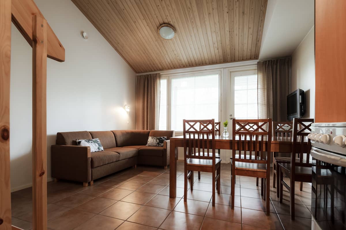 Una sala de estar acogedora con techo de madera, un sofá marrón, una mesa de comedor con sillas y una zona de cocina con estufa.