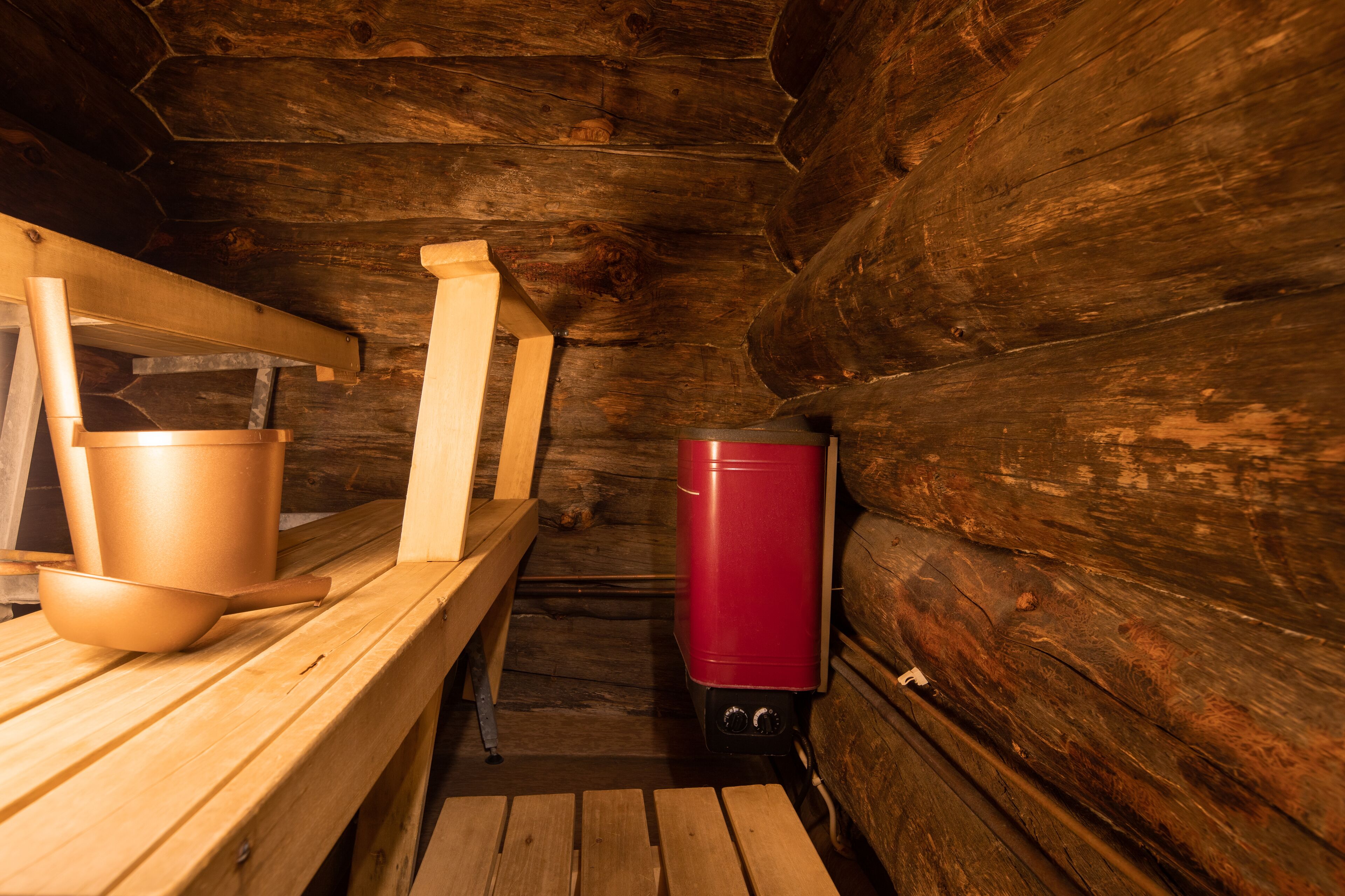 A wooden cabin interior with a bench, a red heater, and a metal bucket.