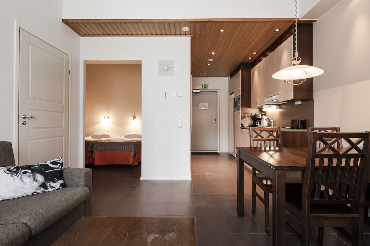 Una habitación de hotel moderna con techo de madera, un área de comedor con mesa y sillas de madera, un sofá con un cojín estampado y una cama con manta roja.